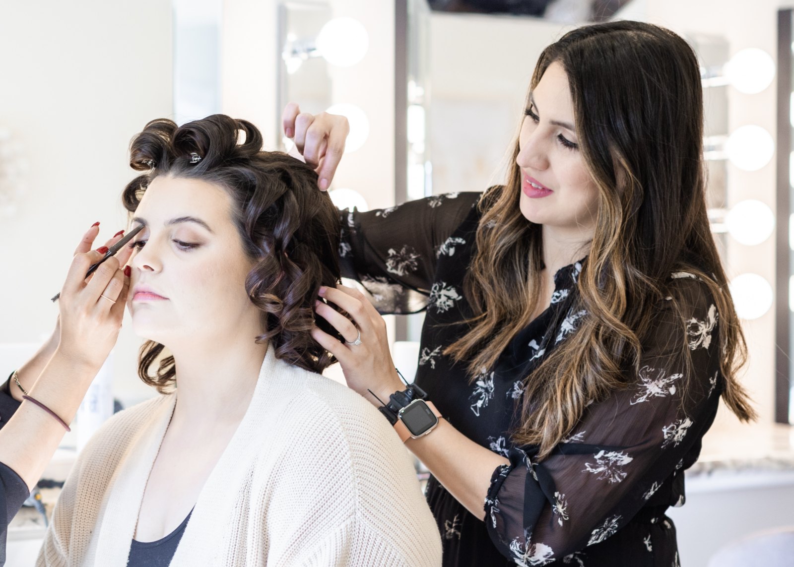 A wedding morning in progress: one artist pins the client’s curls while another applies her eye makeup.